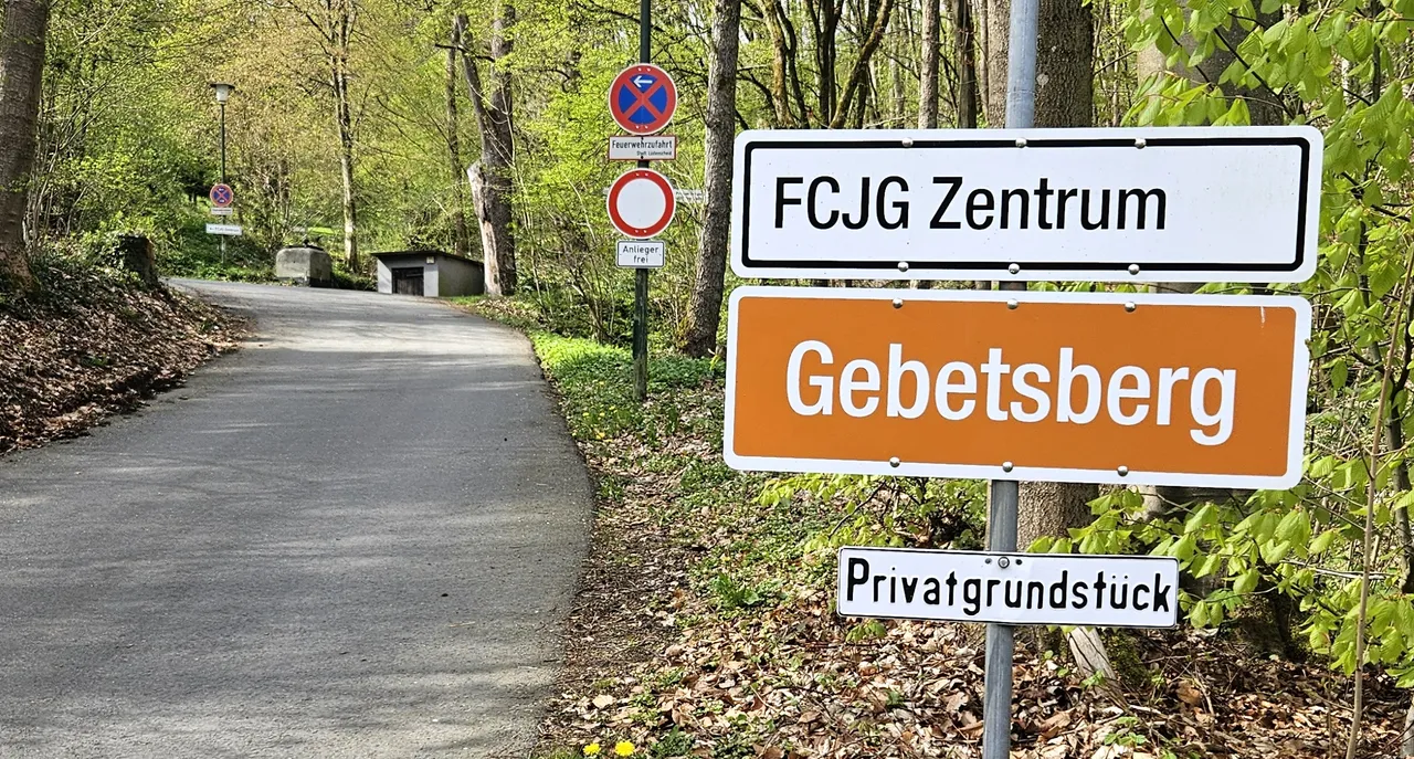 Schilder weisen auf das „FCJG Zentrum“ und „Gebetsberg“ hin, darunter ein Hinweis auf ein Privatgrundstück. Im Hintergrund eine Straße, die sich zwischen Bäumen bergauf windet.