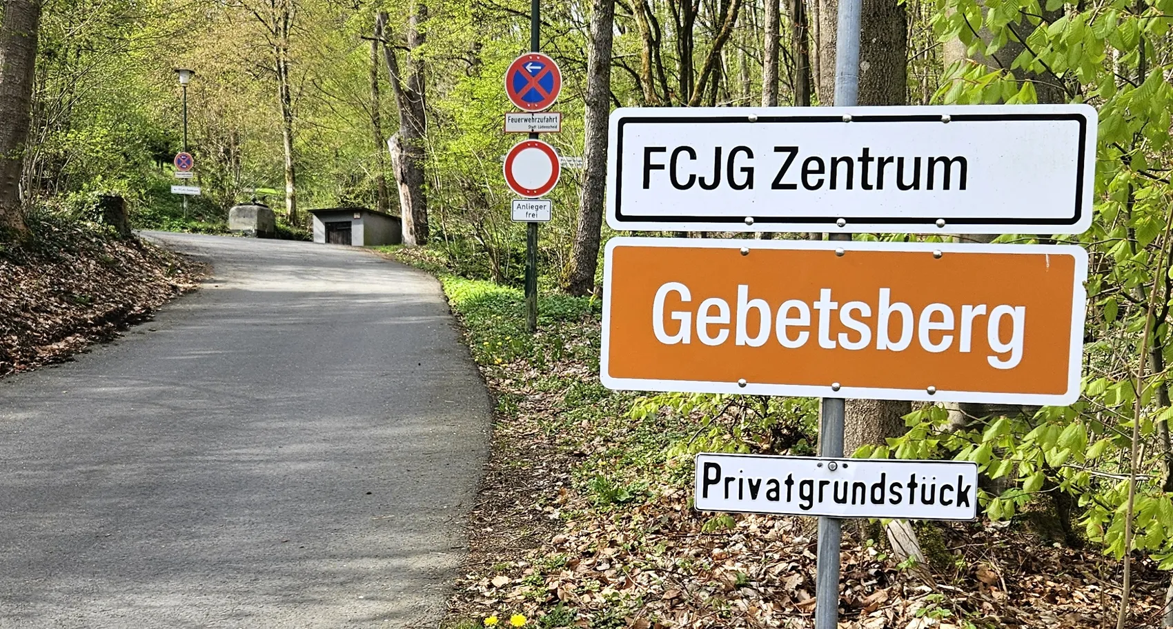Schilder weisen auf das „FCJG Zentrum“ und „Gebetsberg“ hin, darunter ein Hinweis auf ein Privatgrundstück. Im Hintergrund eine Straße, die sich zwischen Bäumen bergauf windet.