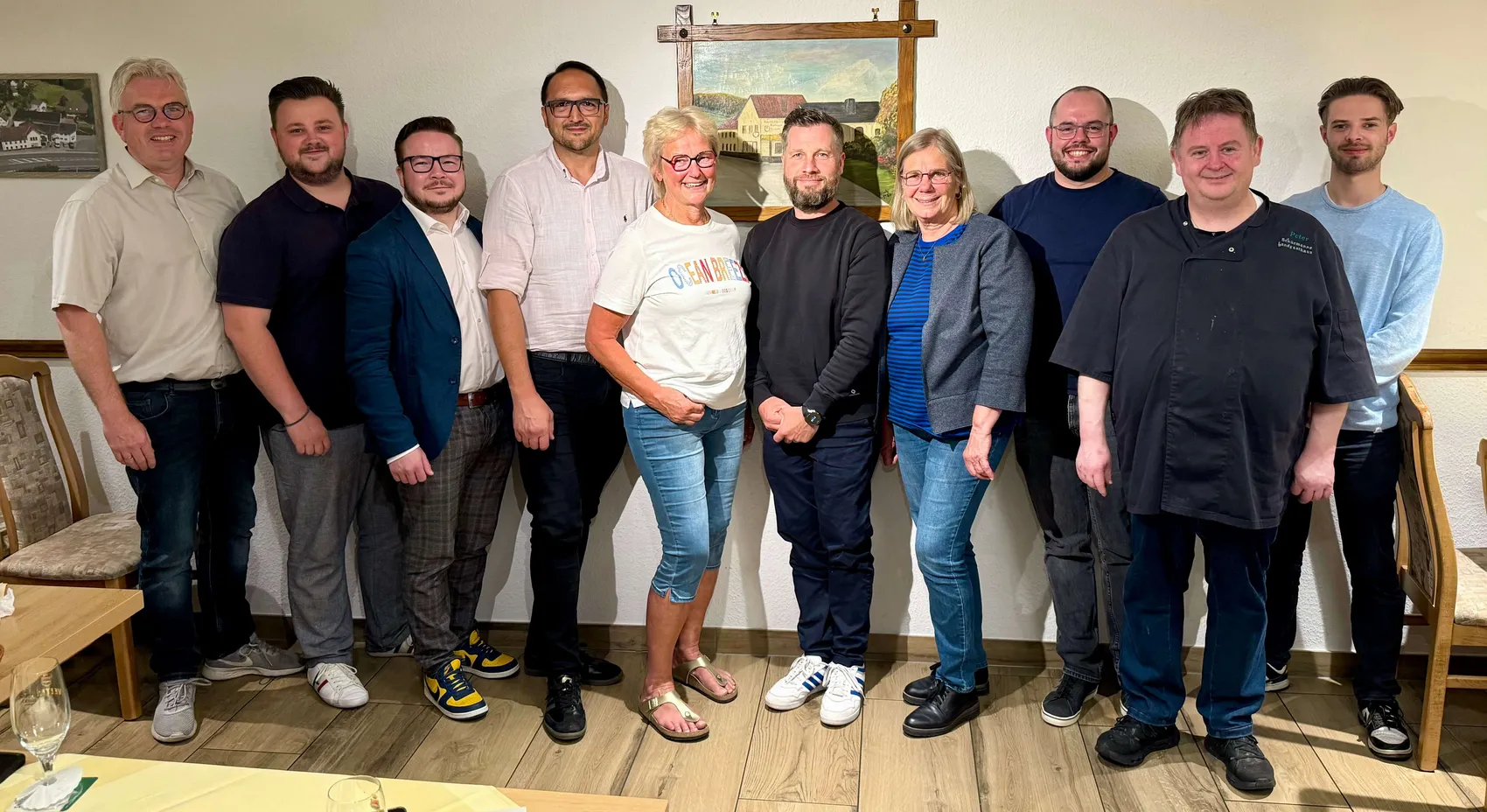 Neun Personen stehen in einem Raum mit Holztäfelung zusammen. Im Zentrum steht eine Frau mit hellen Haaren und Jeans. Die anderen Personen sind unterschiedlich gekleidet, einige tragen Anzüge, andere Freizeitkleidung. Ein Mann rechts trägt eine Kochjacke.