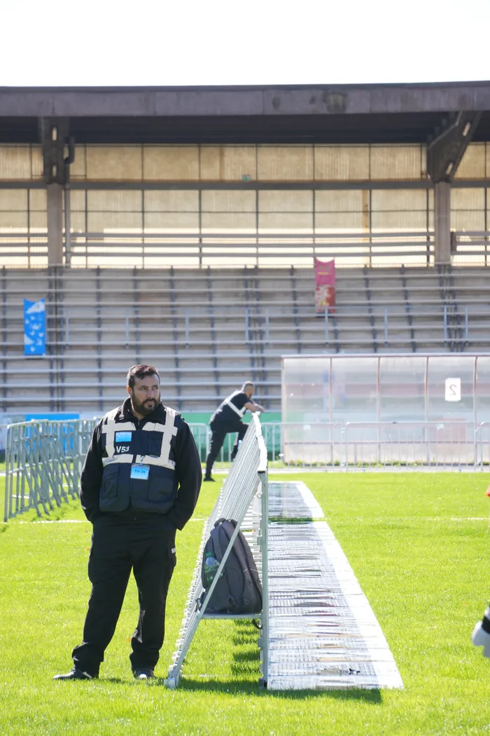 Ein Sicherheitsmitarbeiter in schwarzer Uniform und Weste steht auf einer Rasenfläche vor leeren Stadiontribünen.