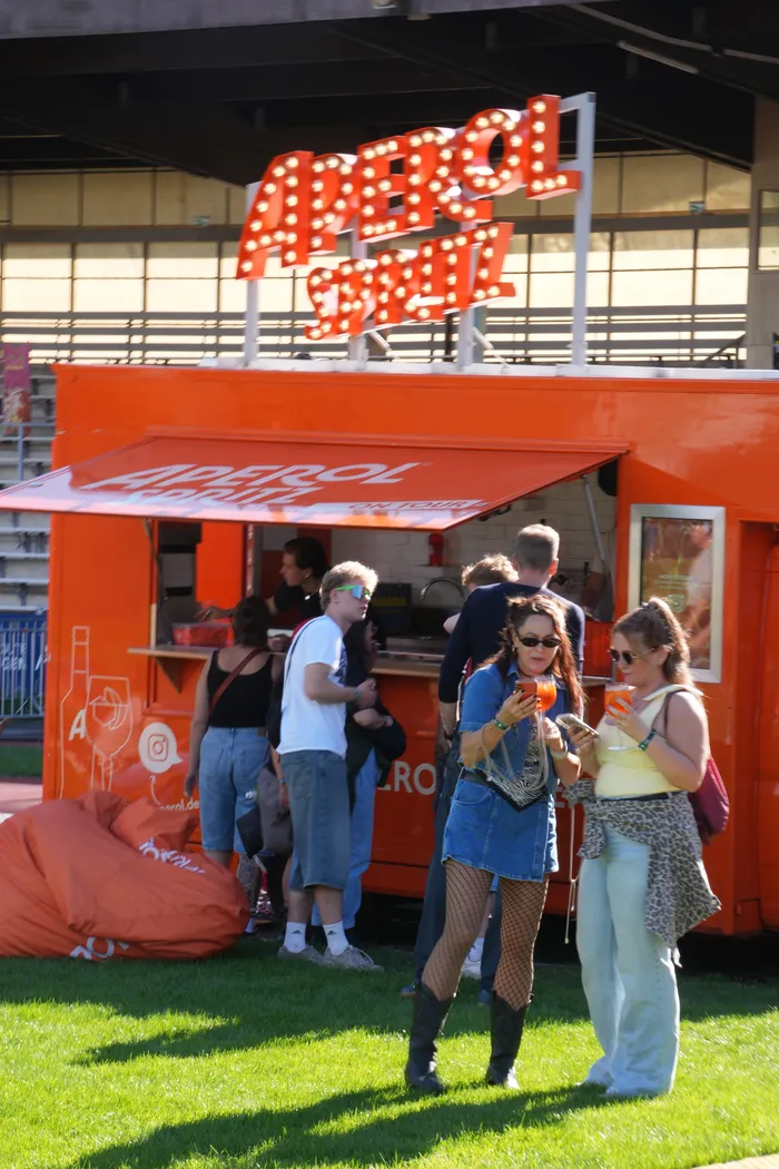 Menschen stehen vor einem orangefarbenen Aperol Spritz Stand mit beleuchtetem Schriftzug auf dem Bautz-Festival.