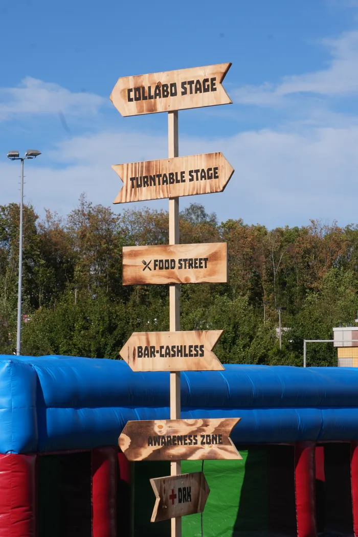 Ein hölzerner Wegweiser auf dem Bautz-Festival mit Richtungen zu der Collabo Stage, Turntable Stage, Food Street, Bar-Cashless, Awareness Zone und DRK.