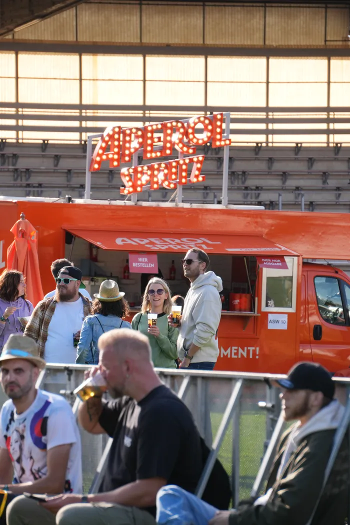 Ein orangefarbener Aperol Spritz Stand mit leuchtendem Schriftzug vor einer Tribüne, umgeben von Menschen auf dem Bautz-Festival.