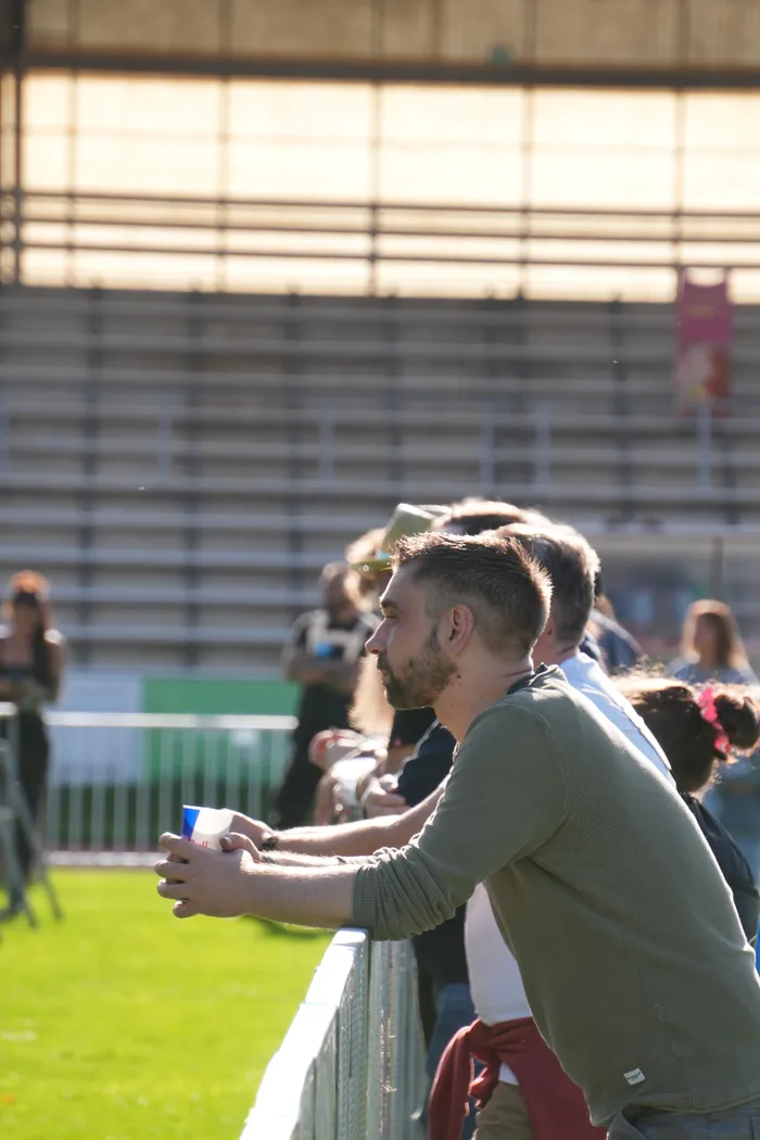 Ein bärtiger Mann in einem grünen Shirt lehnt an einem Metallzaun und hält einen Becher, im Hintergrund sind Tribünen zu sehen.