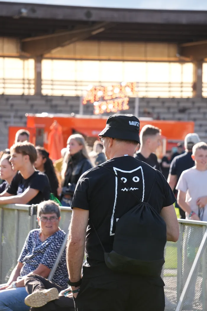 Ein Mann mit schwarzem Bucket-Hat mit der Aufschrift 'BAUTZ' steht mit dem Rücken zur Kamera bei einem Sommerfestival.