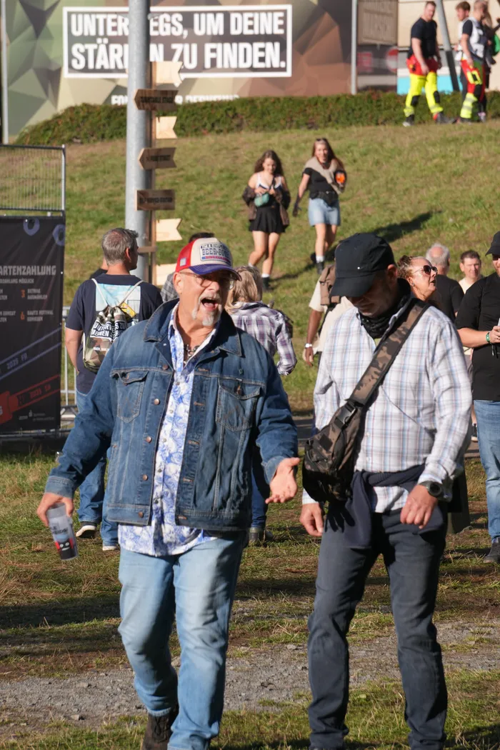 Ein Mann mit roter Kappe und Jeansjacke geht mit offenem Mund über eine Wiese bei einem sonnigen Sommerfest.