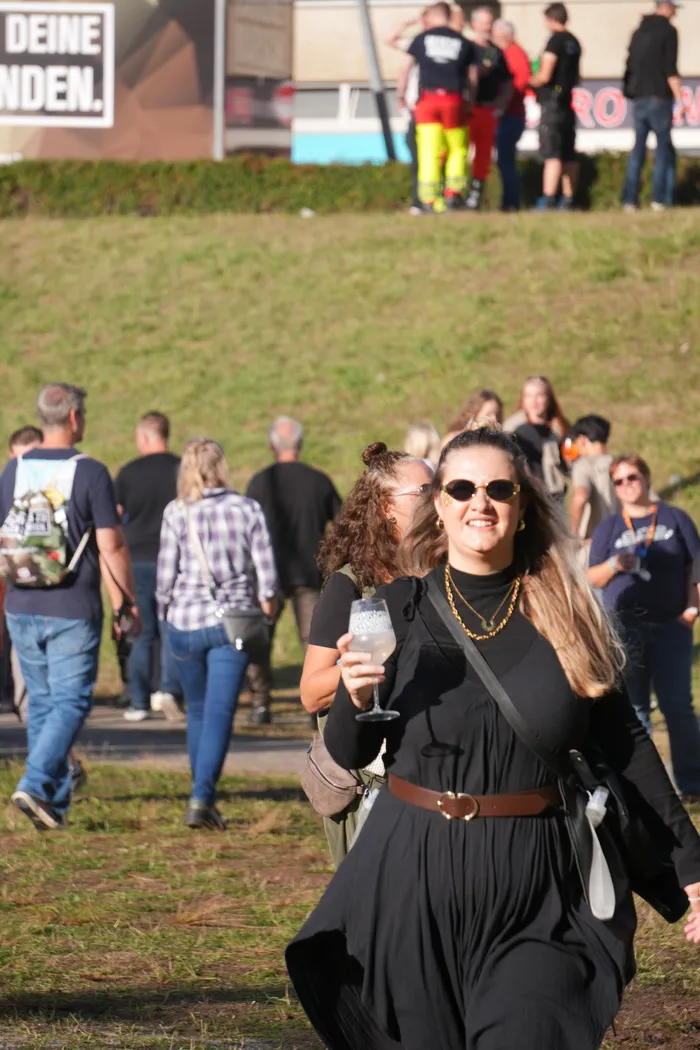 Eine lächelnde Frau in einem schwarzen Kleid geht mit einem Glas Weißwein über eine Wiese bei der Veranstaltung Bautz 2026.