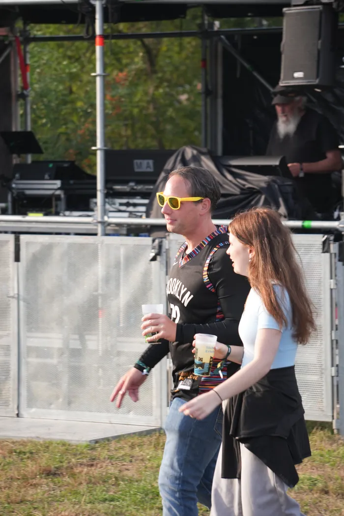 Ein Mann mit gelber Sonnenbrille und eine Frau mit Getränken gehen vor einer Festivalbühne bei Bautz 2026.