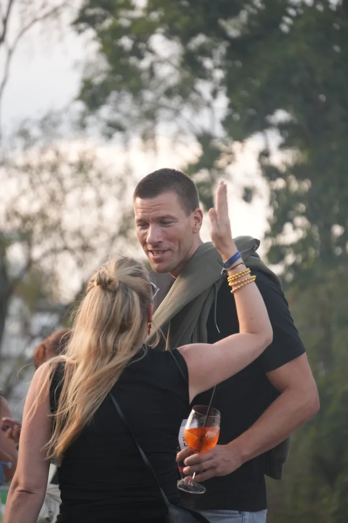 Ein lächelnder Mann hält ein Glas mit einem orangefarbenen Getränk und unterhält sich mit einer Frau, die den Arm hebt.