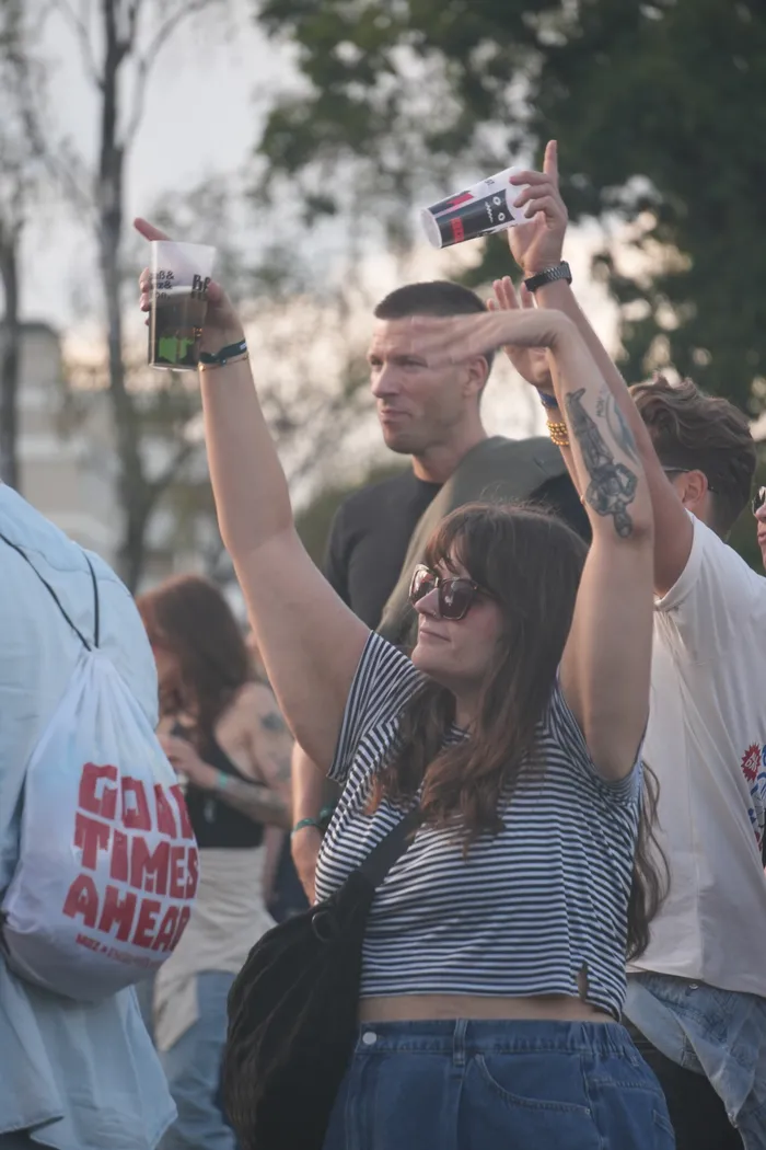 Eine Frau mit Sonnenbrille und gestreiftem Shirt hebt die Arme hoch, während Menschen im Hintergrund bei einem Sommerfest feiern.