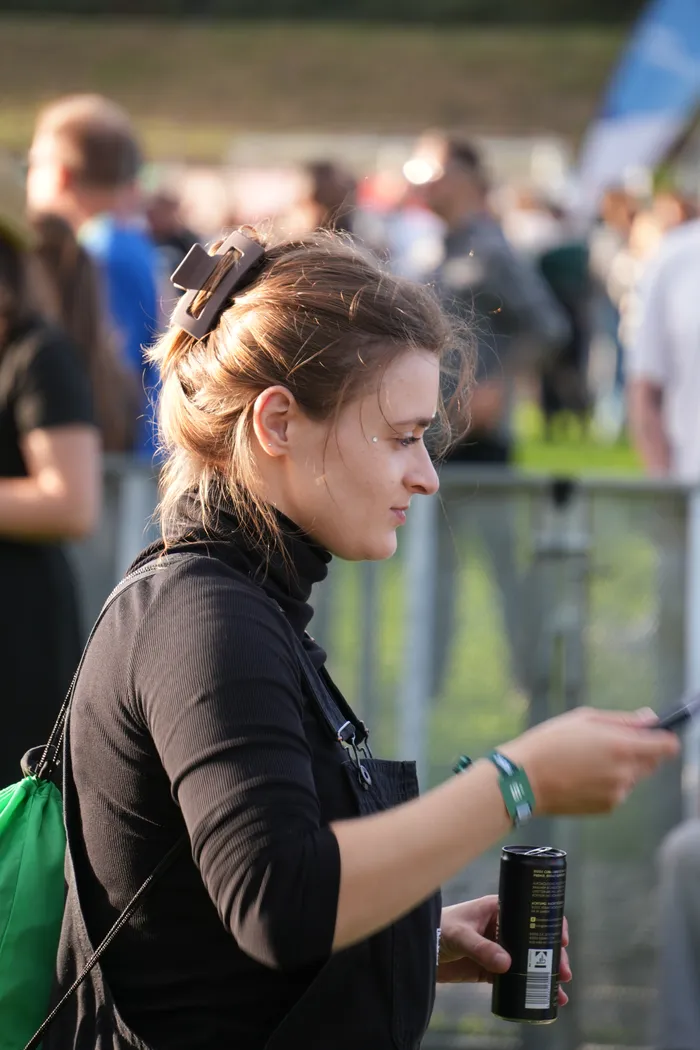 Eine junge Frau blickt auf ihr Smartphone und hält eine schwarze Dose, während im Hintergrund eine Menschenmenge zu sehen ist.