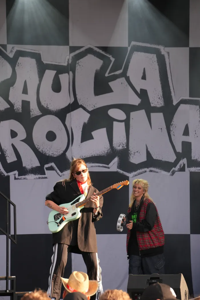 Eine Musikerin mit Sonnenbrille spielt eine hellblaue E-Gitarre vor einem schwarz-weiß karierten Banner mit Graffiti-Schriftzug auf einer Bühne.