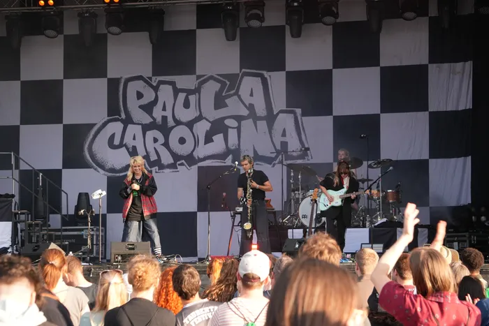 Die Band Paula Carolina tritt auf einer Bühne mit schwarz-weiß kariertem Hintergrund vor einem Publikum auf.