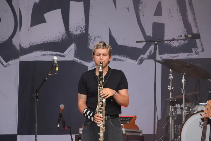 Ein Mann in einem schwarzen T-Shirt spielt Saxophon auf einer Bühne vor einem Hintergrund mit großen grafischen Elementen.