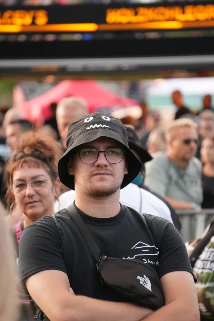 Ein Mann mit Brille und einem schwarzen Bucket Hat mit Augen steht mit verschränkten Armen in einer Menschenmenge in einem Stadion.
