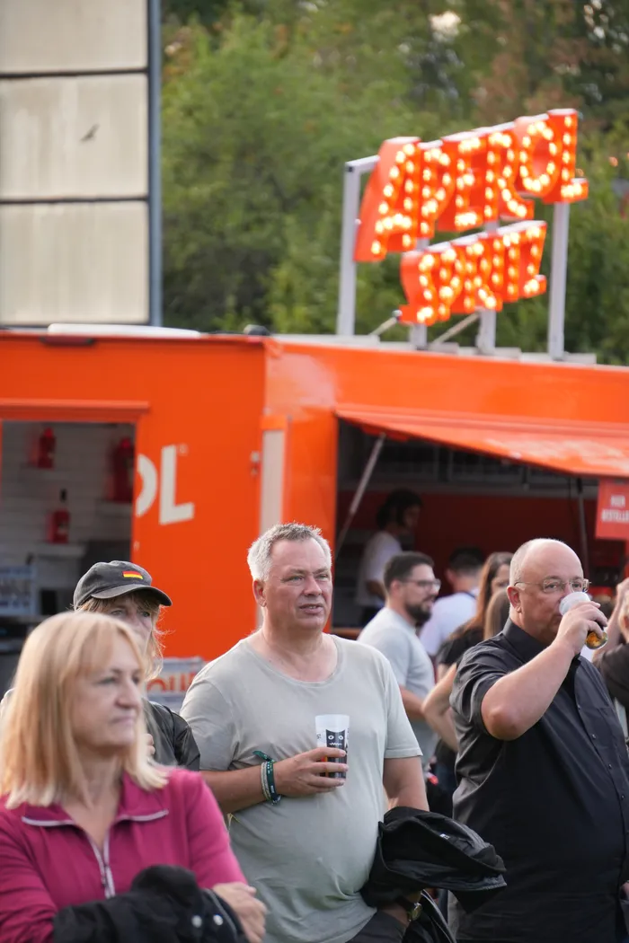 Menschen stehen vor einem orangefarbenen Verkaufsstand mit einem leuchtenden Aperol Spritz Schriftzug.