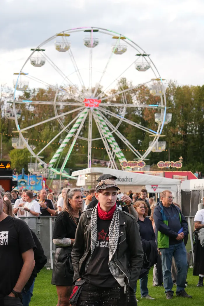 Eine Person in schwarzer Lederjacke und gemustertem Schal steht vor einer Menschenmenge und einem großen Riesenrad.