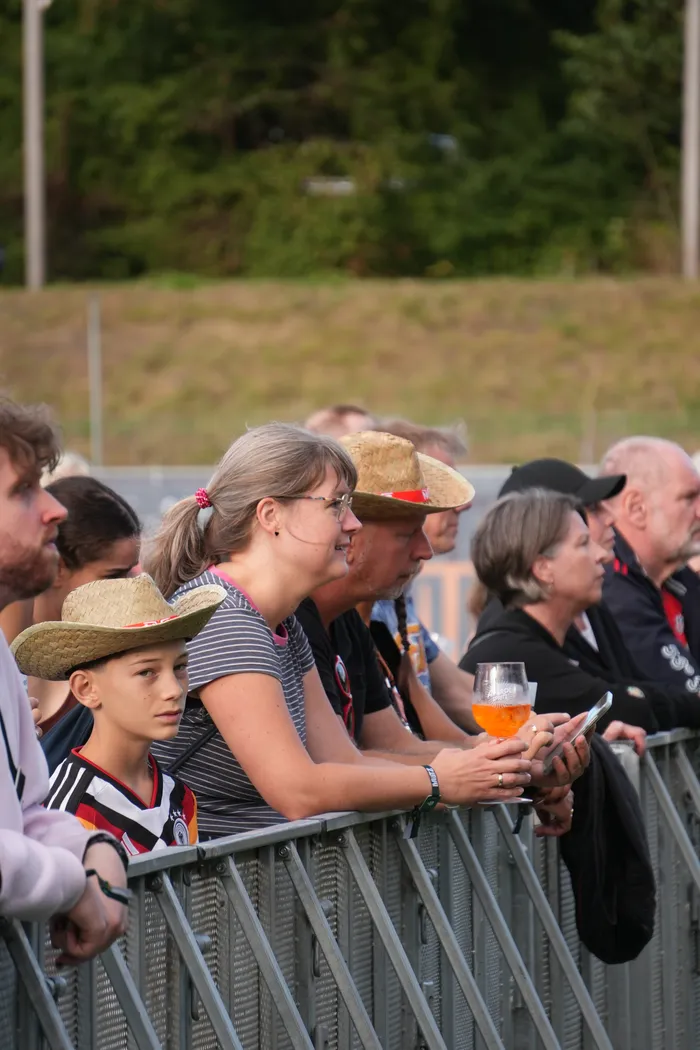 Zuschauer stehen hinter einer Metallabsperrung in einem Stadion. Im Vordergrund sind ein Junge mit Strohhut und eine Frau mit Brille zu sehen.