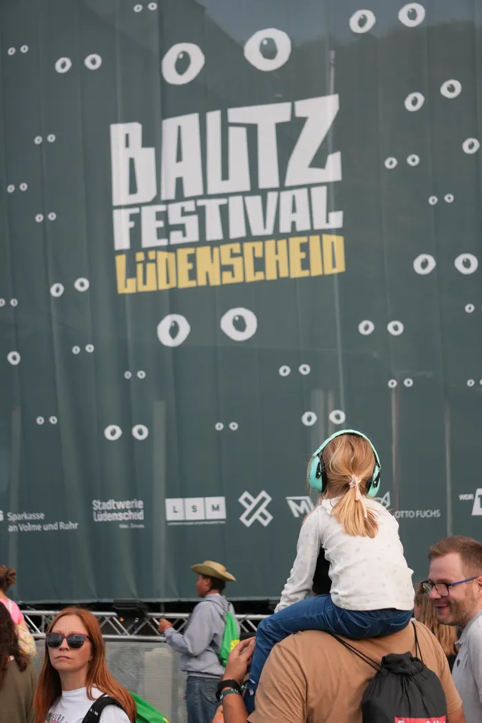 Ein Kind mit hellblauen Kopfhörern sitzt auf den Schultern einer Person vor einem Banner des Baltz Festivals Lüdenscheid.