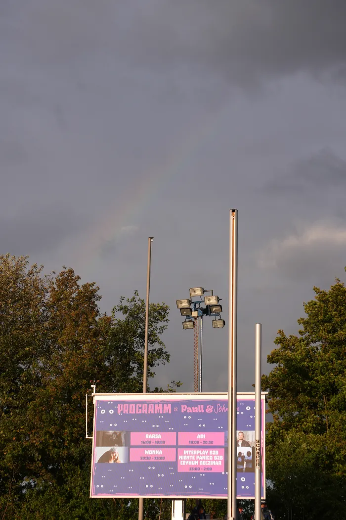 Ein lila Banner mit dem Programm von Paul & Söhne vor Bäumen, einem Lichtmast und einem Regenbogen am bewölkten Himmel.