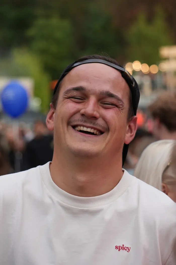 Ein lachender Mann mit Glitzer im Gesicht trägt ein weißes T-Shirt mit der Aufschrift „spicy“ und eine schwarze Kappe.
