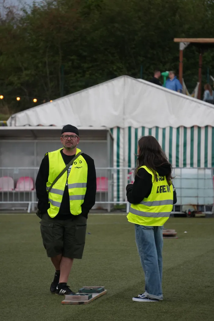 Zwei Personen in neongelben Warnwesten stehen auf einer Rasenfläche vor einem gestreiften Zelt.