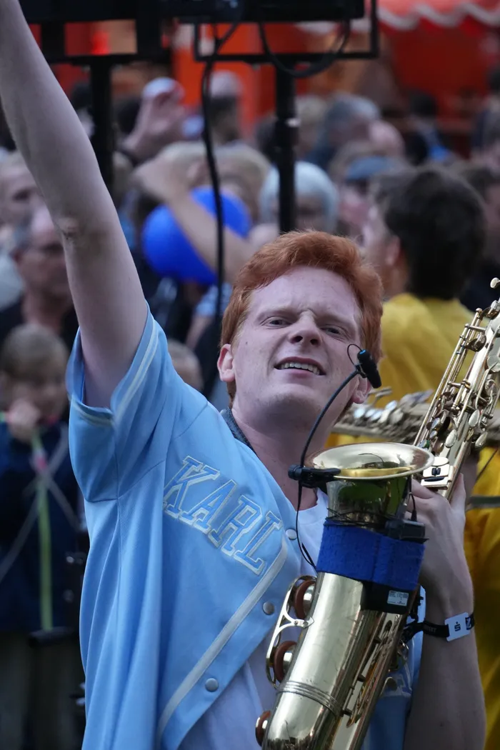 Ein rothaariger Musiker in einem hellblauen Shirt mit der Aufschrift „KARL“ spielt Saxophon und hebt einen Arm in einer Menschenmenge.