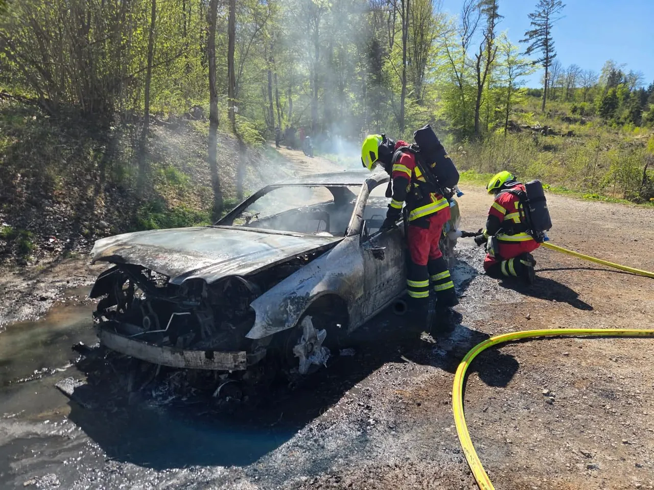 Feuerwehrleute löschen einen völlig ausgebrannten Pkw an einer Landstraße, während Rauch aufsteigt.
