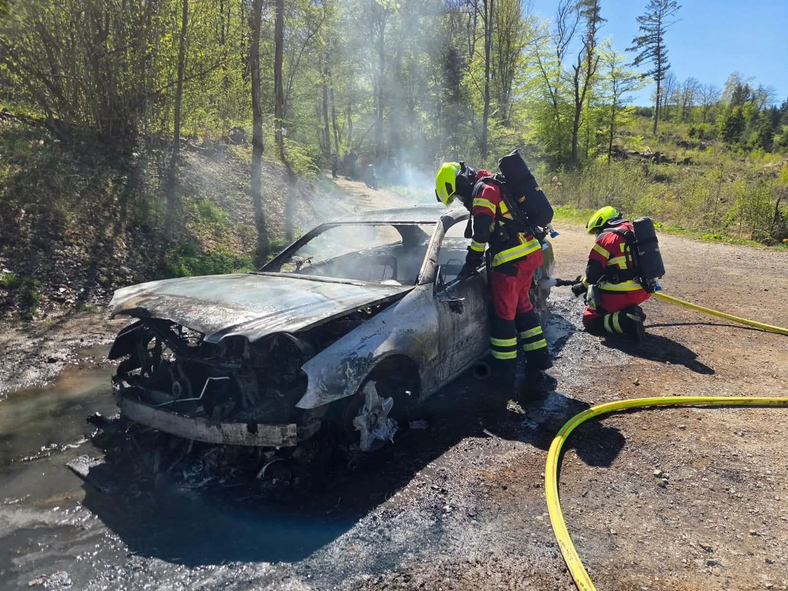 Feuerwehrleute löschen einen völlig ausgebrannten Pkw an einer Landstraße, während Rauch aufsteigt.