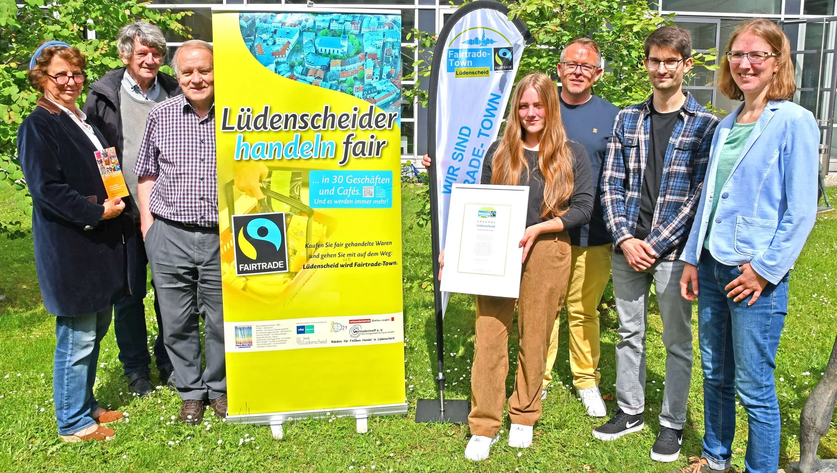 Eine Gruppe von sieben Personen hält ein Schild mit der Aufschrift „Wir sind Fairtrade-Town“ und eine Urkunde. Im Hintergrund ein Banner mit dem Slogan „Lüdenscheider handeln fair“ und dem Fairtrade-Logo.