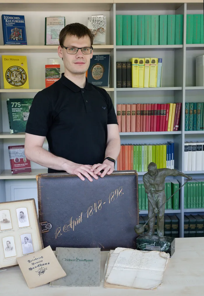 Archivar Lars Lötters steht vor einem Bücherregal und einem geöffneten Koffer mit historischen Dokumenten und Fotografien der Familie Brockhaus. Eine Bronzestatue ist ebenfalls zu sehen.