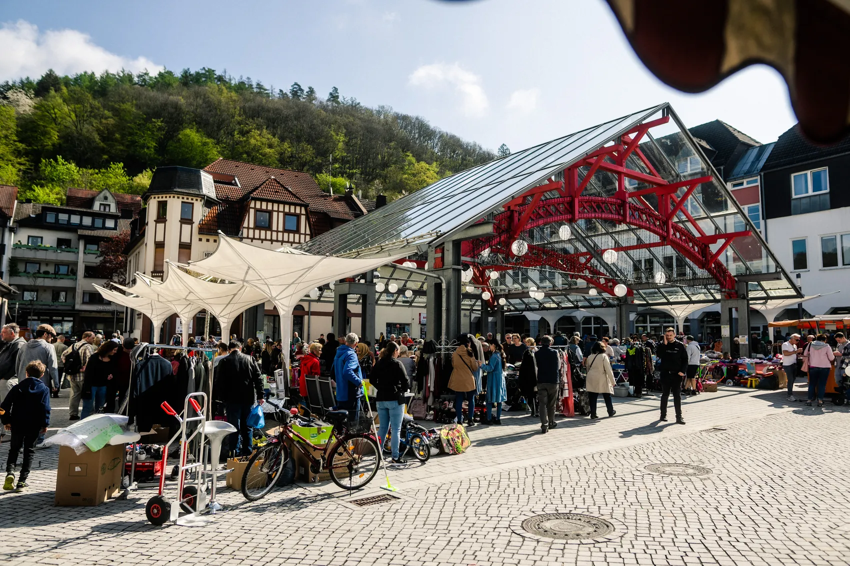 Blick über einen belebten Platz mit Marktständen und Besuchern unter einem großen, modernen Glas- und Stahlkonstruktion. Im Hintergrund sind Fachwerkhäuser und ein bewaldeter Hügel zu sehen.
