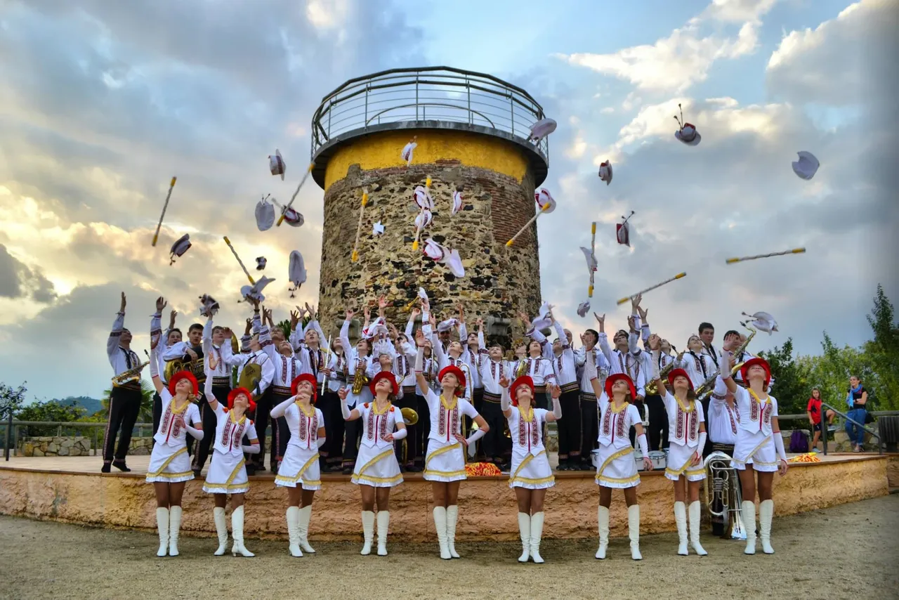 Eine Gruppe von etwa 30 Musikern in historischen Uniformen wirft ihre Hüte in die Luft, vor einem mittelalterlichen Wachturm. Im Hintergrund ein bewölkter Himmel.