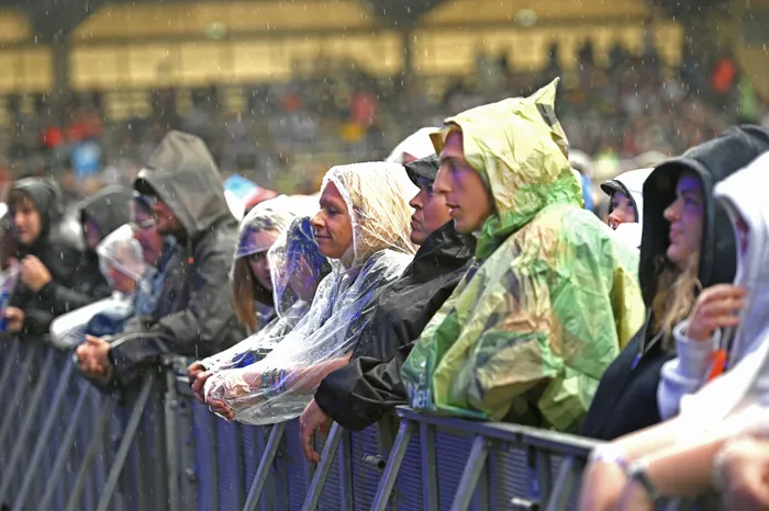 Fans in Regenponchos und Kapuzenjacken stehen bei starkem Regen hinter einer Metallabsperrung.