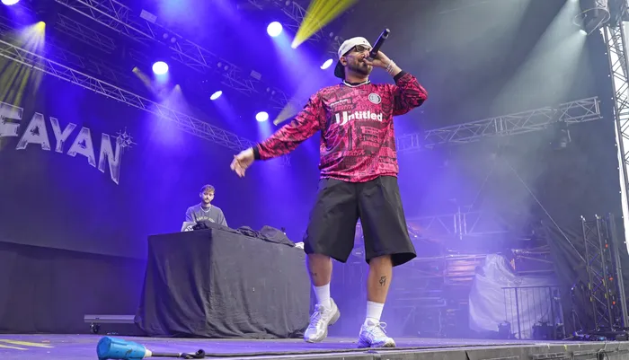 Fayan performt mit einem Mikrofon auf einer Bühne unter blauem und gelbem Scheinwerferlicht. Er trägt ein pink-schwarzes gemustertes Shirt und schwarze Shorts.