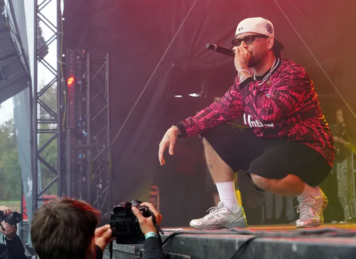 Fayan in einem pink-schwarzen gemusterten Oberteil und weißer Kappe singt hockend in ein Mikrofon auf einer Bühne.