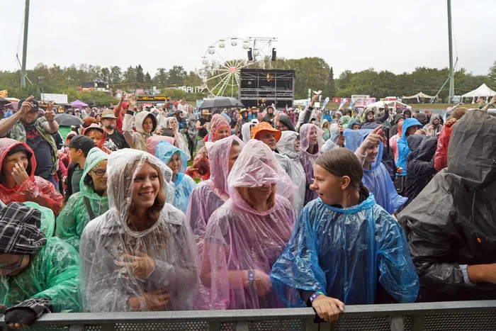 Eine Menschenmenge in bunten Regenponchos blickt bei einem Open-Air-Event auf eine Bühne, im Hintergrund ist ein Riesenrad zu sehen.