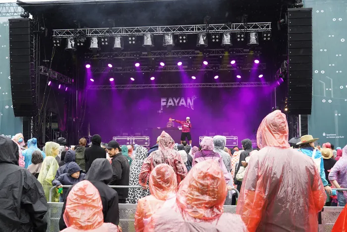 Fayan performt auf einer lila beleuchteten Bühne vor einer Menschenmenge in bunten Regenponchos.