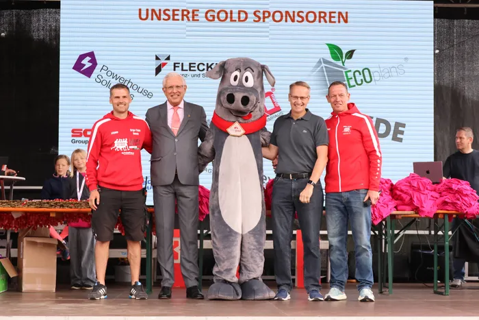 Vier Männer und ein graues Maskottchen posieren auf einer Bühne vor einer Sponsorenwand.