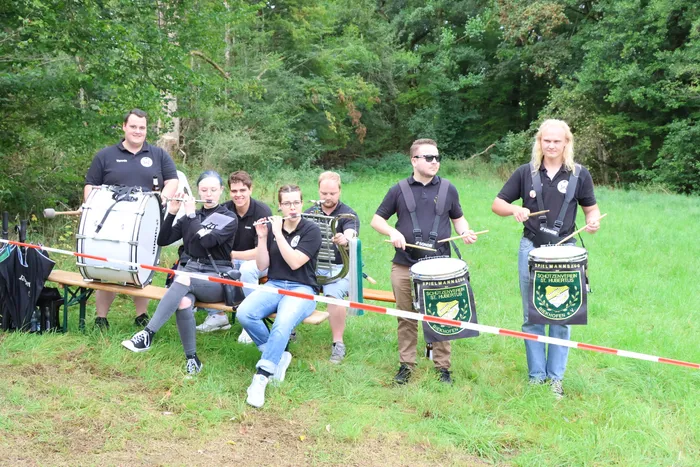 Eine Gruppe von Musikern spielt Trommeln und Blasinstrumente auf einer Wiese vor einem Waldrand.