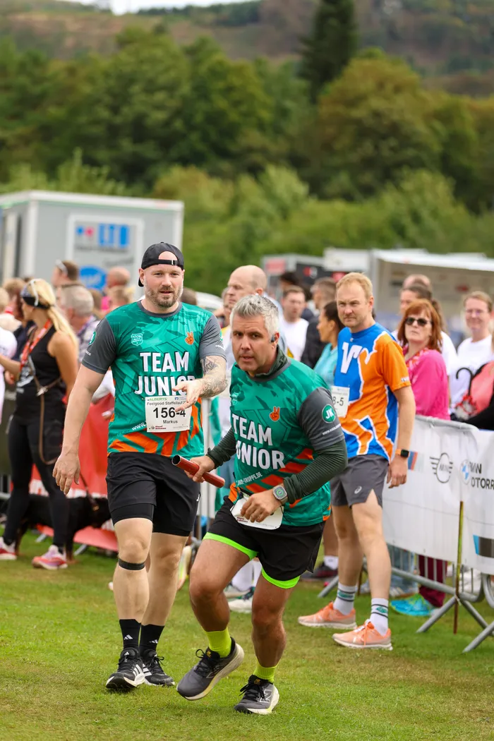Zwei Männer in grünen „TEAM JUNIOR“-Shirts beim Feelgood-Staffellauf in Hüinghausen, einer hält einen roten Staffelstab.