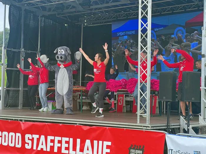 Menschen in roten T-Shirts und ein graues Maskottchen winken auf einer Bühne beim Feelgood-Staffellauf einer Menschenmenge zu.