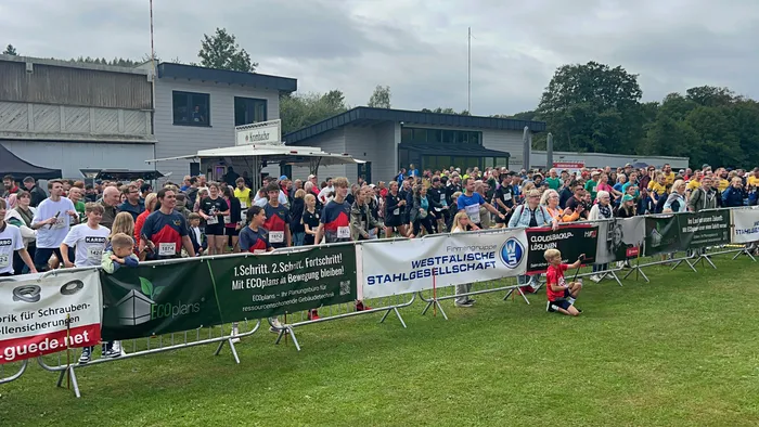 Eine große Gruppe von Menschen in Sportkleidung versammelt sich bei einem Staffellauf auf einer Wiese hinter Sponsorenbannern.