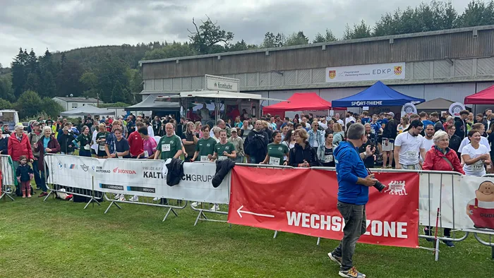 Teilnehmer eines Staffellaufs stehen hinter Absperrungen vor Zelten; ein rotes Banner im Vordergrund markiert die „WECHSZONE“.