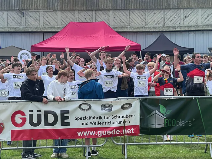 Eine große Gruppe junger Teilnehmer in weißen T-Shirts mit Startnummern hebt die Hände bei einem Staffellauf.