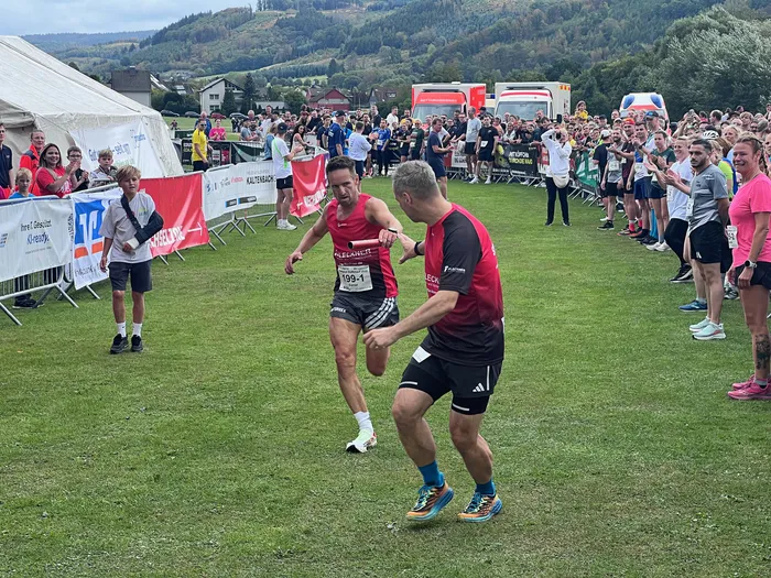 Zwei Männer in roter Sportkleidung laufen bei einem Staffellauf über eine Wiese vor einer Zuschauermenge.