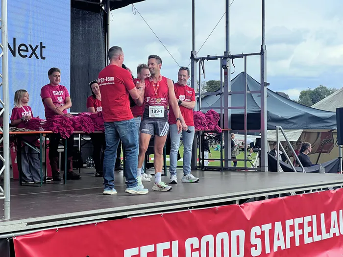 Ein Läufer mit Startnummer steht auf einer Bühne und wird von Teammitgliedern in roten T-Shirts empfangen.