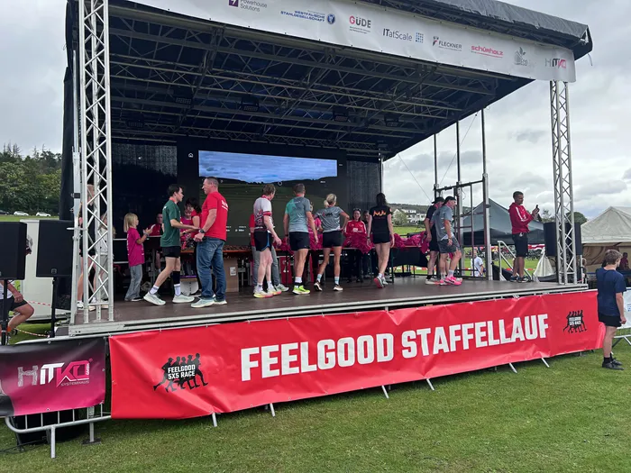 Eine Gruppe von Menschen steht auf einer Bühne; im Vordergrund ist ein rotes Banner mit der Aufschrift „FEELGOOD STAFFELLAUF“ zu sehen.