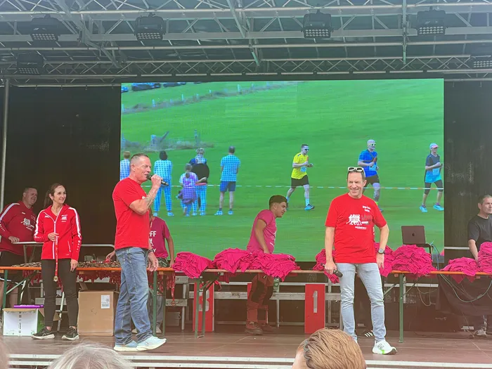 Personen in roten T-Shirts auf einer Bühne vor einer Leinwand, die Läufer bei einem Staffellauf zeigt.