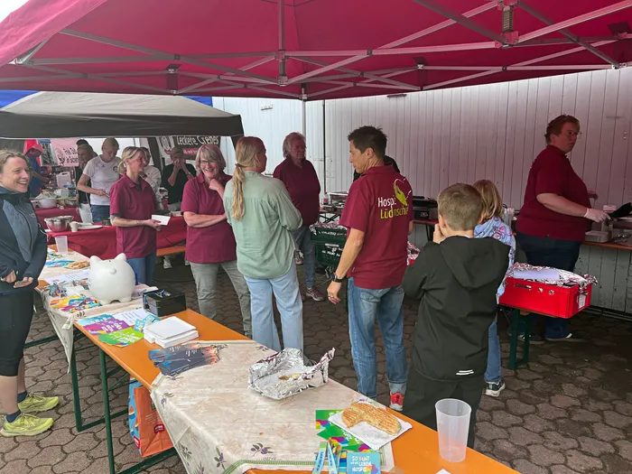 Menschen stehen unter einem roten Zelt an Tischen mit Speisen, Getränken und einer Spardose.